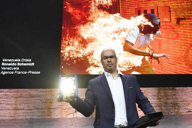 Venezualan AFP photographer Ronaldo Schemidt holds his trophy in Amsterdam on April 12, 2018 during the 2018 World Press Photo (WPP) award ceremony after being awarded with both picture of the Year 2018 award and 1st prize in the Spot News Singles category for the picture projected above taken on May 3, 2017 of a demonstrator catching fire during clashes with riot police within a protest against Venezuelan President in Caracas. / AFP PHOTO / EMMANUEL DUNAND